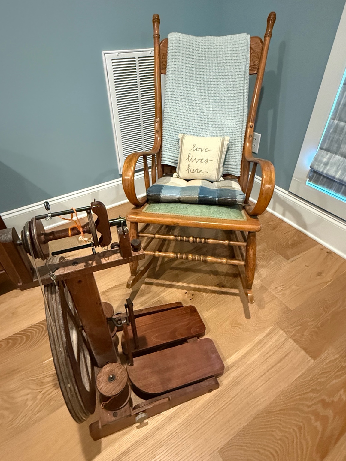 Antique spinning wheel beside a wooden rocking chair with a cozy blanket and a pillow that says Love Lives Here.